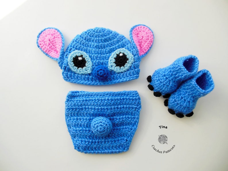 May include: A blue crocheted baby hat, diaper cover, and booties set featuring a Stitch character design. The hat has pink ears, black eyes, and a blue nose. The booties have black toes.