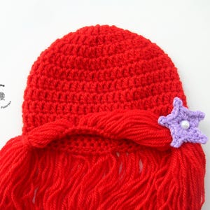 May include: Red crocheted hat with long red yarn strands, resembling hair, and a purple star-shaped embellishment with a white pearl in the center.