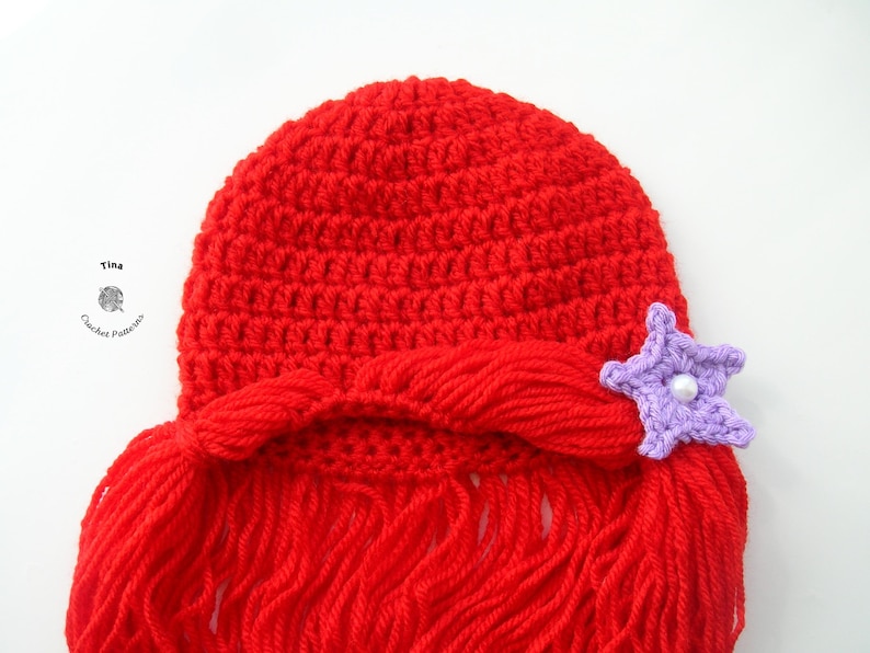 May include: Red crocheted hat with long red yarn strands, resembling hair, and a purple star-shaped embellishment with a white pearl in the center.
