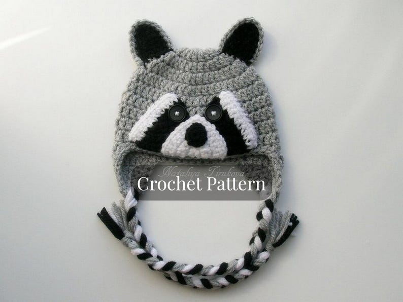 May include: Gray crocheted raccoon hat with black and white details. The hat has a long, braided tail and the text "Crochet Pattern" is printed on the front.