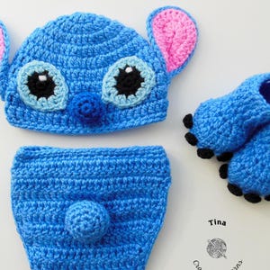 May include: Blue crocheted baby hat, diaper cover, and booties with black eyes and a pink nose. The hat has large pink ears. The booties have black accents. The set is labeled "Tina Crochet Patterns".