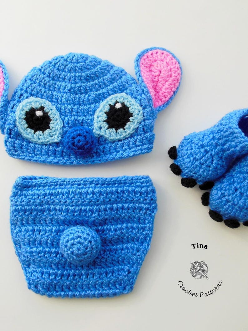 May include: Blue crocheted baby hat, diaper cover, and booties with black eyes and a pink nose. The hat has large pink ears. The booties have black accents. The set is labeled "Tina Crochet Patterns".