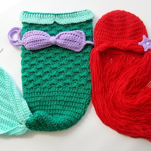 May include: A crocheted mermaid costume for a baby, featuring a green and turquoise tail, a purple bow, and a red crocheted wig with a purple starfish.