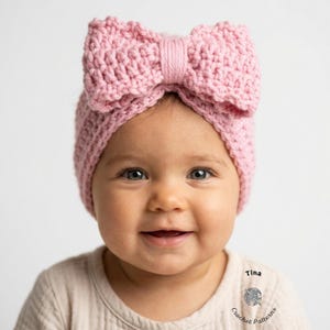 May include: A baby wearing a light pink, crocheted headband with a large bow. The headband is made of a thick yarn and has a textured appearance. The baby is smiling and wearing a beige shirt.