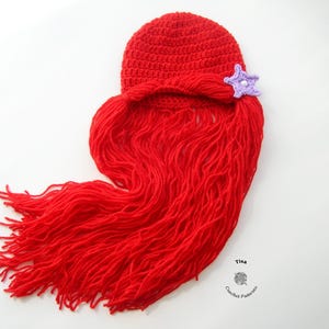 May include: Red crocheted hat with a long red yarn tail, resembling a mermaid's hair. A purple crocheted star is attached to the hat.