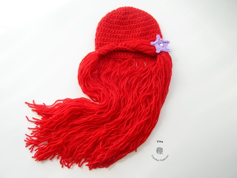 May include: Red crocheted hat with a long red yarn tail, resembling a mermaid's hair. A purple crocheted star is attached to the hat.