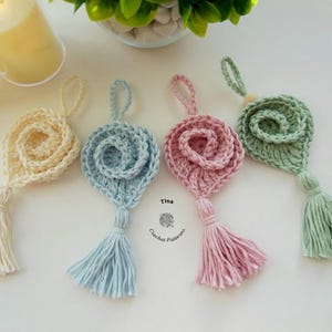 May include: Four crocheted flower ornaments with tassels in different colors: cream, light blue, pink, and green. The ornaments are hanging from a white surface. The text "Tina Crochet Patterns" is visible in the center of the image.
