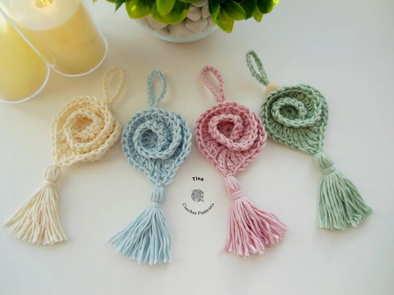 May include: Four crocheted flower ornaments with tassels in different colors: cream, light blue, pink, and green. The ornaments are hanging from a white surface. The text "Tina Crochet Patterns" is visible in the center of the image.