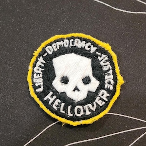 May include: A circular embroidered patch featuring a white skull design centered on a black background. The border is bright yellow. The text "LIBERTY - DEMOCRACY - HELLDIVER" is stitched in white capital letters around the central skull motif.