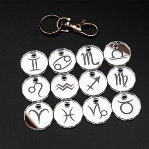 May include: Twelve silver-coloured keychains with zodiac symbols. Each keychain is a round, mirrored disc with a different zodiac symbol etched on it. The keychains are arranged in three rows of four. A silver keychain ring is included.