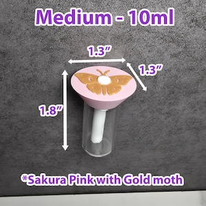 May include: A clear glass bottle with a pink lid featuring a gold moth design. The bottle is labeled "Medium - 10ml" and measures 4.57 cm in height and 3.3 cm in width. The lid is labeled "Sakura Pink with Gold moth".