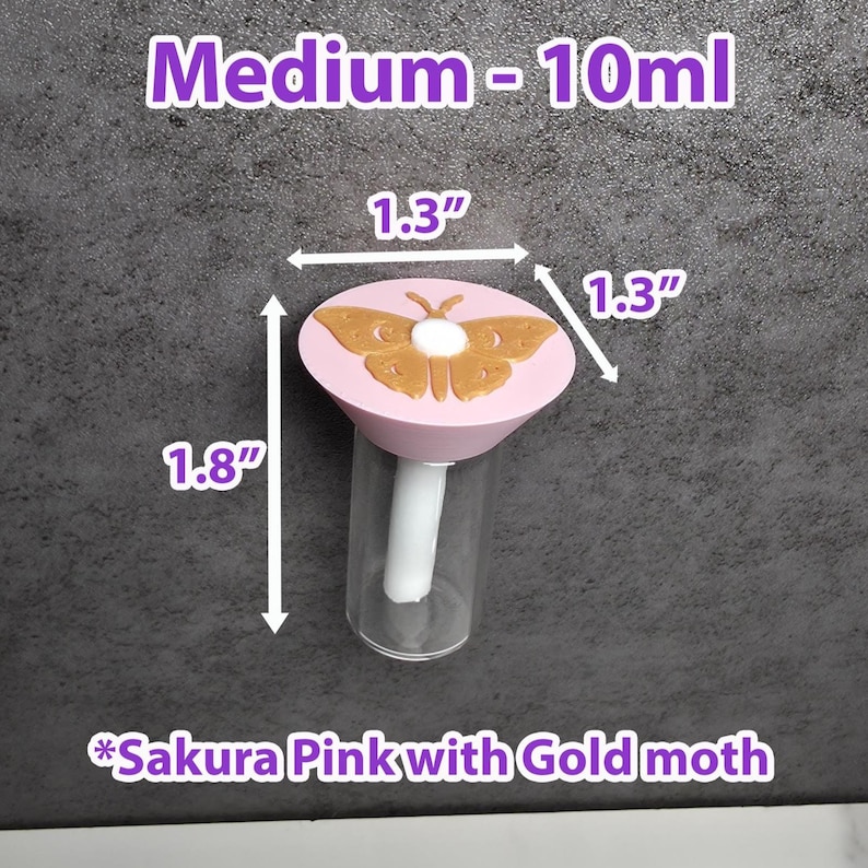May include: A clear glass bottle with a pink lid featuring a gold moth design. The bottle is labeled "Medium - 10ml" and measures 4.57 cm in height and 3.3 cm in width. The lid is labeled "Sakura Pink with Gold moth".