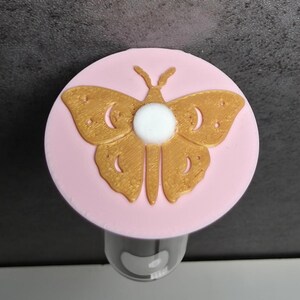 May include: A bottle stopper with a gold butterfly design on a pink disc. The butterfly has detailed wings and a white central element. The stopper is designed to fit a bottle, adding a decorative touch.