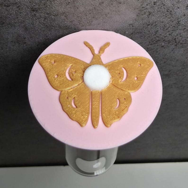 May include: A bottle stopper with a gold butterfly design on a pink disc. The butterfly has detailed wings and a white central element. The stopper is designed to fit a bottle, adding a decorative touch.