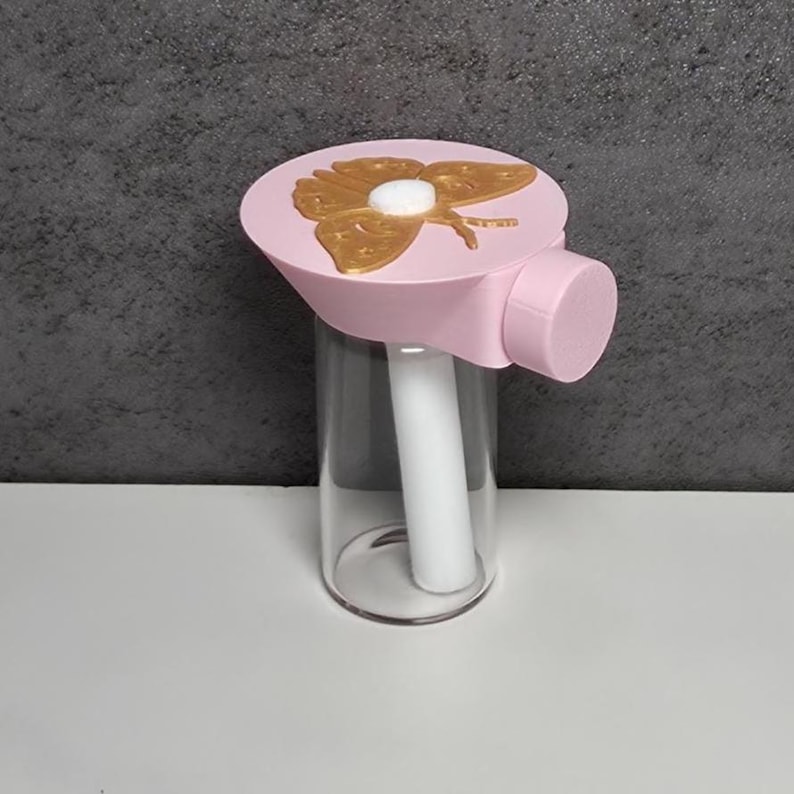 May include: A pink bottle stopper with a gold butterfly design. The stopper is attached to a clear glass bottle with a white interior component. The butterfly design is gold with a white center. The stopper is a light pink colour.
