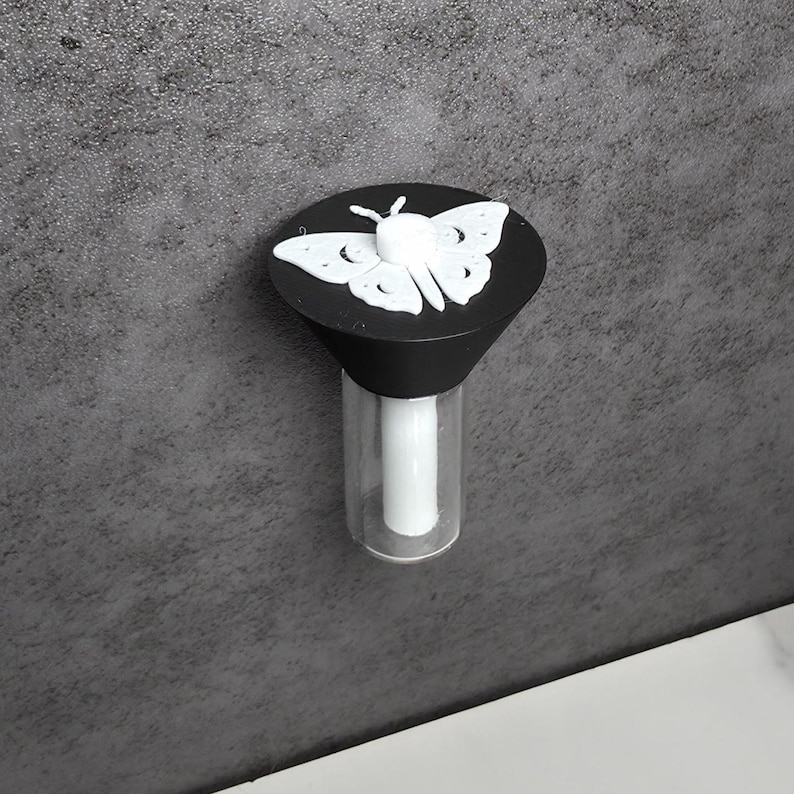 May include: A black bottle stopper featuring a white butterfly design. The butterfly is centrally placed on the stopper's top. The stopper has a clear glass base with a white cylindrical insert. The background is a textured grey surface.