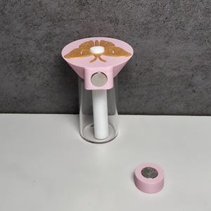 May include: A pink and gold butterfly-topped bottle stopper with a clear glass bottle. The stopper has a magnetic closure and a separate pink magnetic disc. The butterfly design is gold and the bottle is clear.