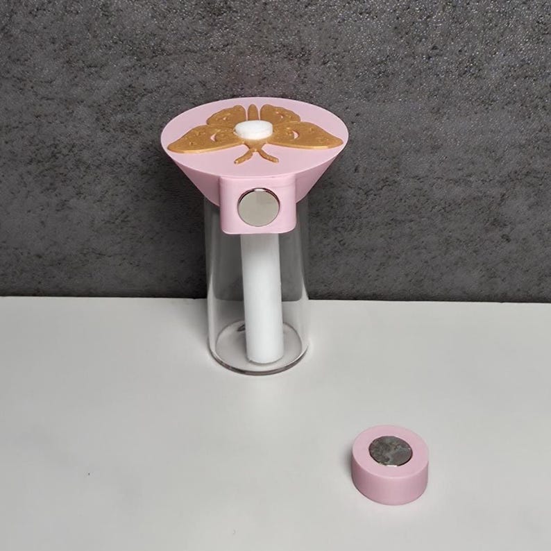 May include: A pink and gold butterfly-topped bottle stopper with a clear glass bottle. The stopper has a magnetic closure and a separate pink magnetic disc. The butterfly design is gold and the bottle is clear.