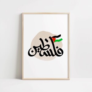Könnte beinhalten: Ein weißer gerahmter Druck mit einem beigefarbenen Aquarellhintergrund, der schwarze arabische Kalligraphie zeigt, die "فلسطين" (Palästina) lautet, mit einer palästinensischen Flagge in der oberen rechten Ecke.