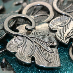 May include: Silver metal charms shaped like leaves with intricate details. The charms have a textured surface and are ideal for jewellery making or crafting.
