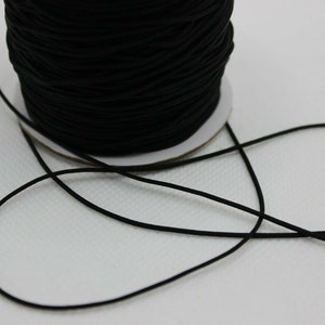May include: A reel of black elastic cord. The cord is tightly wound around a cardboard core. A length of the cord extends from the reel, lying loosely against a white background. The cord appears smooth and flexible.