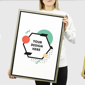 May include: Three people are holding up blank poster frames with black, silver, and gold frames. The posters have a geometric design with a hexagon shape that says "Your Design Here" in black text. The background is a light grey.