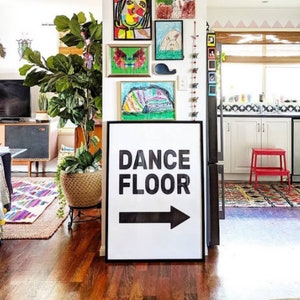 May include: A black and white sign with the words "DANCE FLOOR" and a black arrow pointing to the right. The sign is hanging on a wall in a home with colorful artwork and a potted plant.
