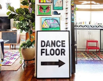 May include: A black and white sign with the words "DANCE FLOOR" and a black arrow pointing to the right. The sign is hanging on a wall in a home with colourful artwork and a potted plant.