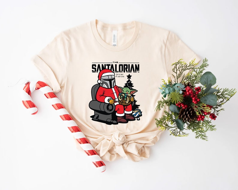 Puede incluir: Una camiseta beige con un gr&aacute;fico de un personaje mandaloriano vestido de Pap&aacute; Noel sosteniendo un peque&ntilde;o alien&iacute;gena verde. El texto "The Santalorian" est&aacute; por encima del gr&aacute;fico. El texto debajo del gr&aacute;fico dice "Did I get it right? It's just a box, right?"