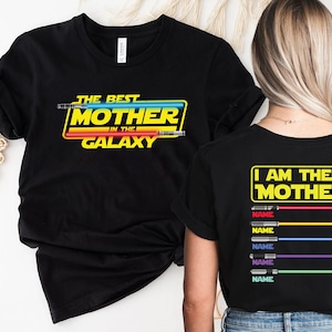 Puede incluir: Dos camisetas negras con un dise&ntilde;o de tem&aacute;tica de Star Wars. La camiseta delantera dice "The Best Mother in the Galaxy" con un gr&aacute;fico de sable de luz. La camiseta trasera dice "I Am Their Mother" con un gr&aacute;fico de sable de luz y espacio para nombres.