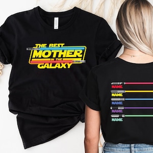 Puede incluir: Camiseta negra con el texto "The Best Mother in the Galaxy" en amarillo y rojo con un gr&aacute;fico de sable de luz. La parte posterior de la camiseta tiene cinco gr&aacute;ficos de sable de luz con la palabra "NAME" debajo de cada uno.