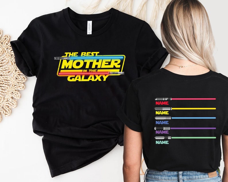 Puede incluir: Camiseta negra con el texto "The Best Mother in the Galaxy" en amarillo y rojo con un gr&aacute;fico de sable de luz. La parte posterior de la camiseta tiene cinco gr&aacute;ficos de sable de luz con la palabra "NAME" debajo de cada uno.