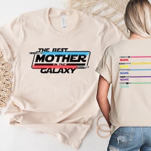 Puede incluir: Una camiseta beige con el texto "The Best Mother in the Galaxy" en negro, rojo y azul. El texto est&aacute; rodeado por un contorno negro y un gr&aacute;fico de sable de luz azul y rojo. La parte posterior de la camiseta tiene cinco sables de luz de diferentes colores con la palabra "NAME" impresa debajo de cada uno.