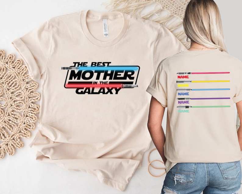 Puede incluir: Una camiseta beige con el texto "The Best Mother in the Galaxy" en negro, rojo y azul. El texto est&aacute; rodeado por un contorno negro y un gr&aacute;fico de sable de luz azul y rojo. La parte posterior de la camiseta tiene cinco sables de luz de diferentes colores con la palabra "NAME" impresa debajo de cada uno.
