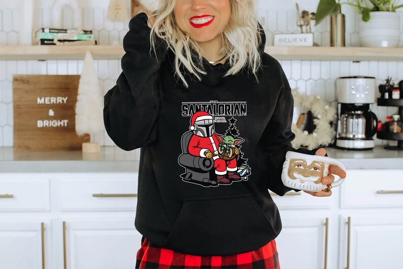 Puede incluir: Sudadera con capucha negra con un gr&aacute;fico de un Santa Mandalorian sosteniendo un beb&eacute; Yoda. El texto en la sudadera dice "The Santaltorian".