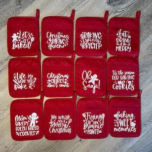 May include: Red quilted oven mitts with white text. Phrases include "Let's Get Baked", "Christmas Baking Queen", "Baking Spirits Bright", and "Eat Drink & Be Merry". Other mitts say "Life is What You Bake It".