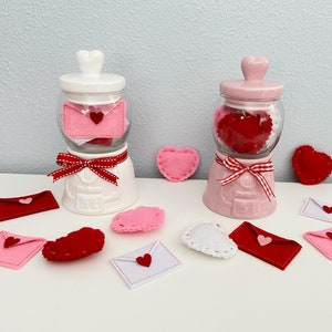 May include: Two white ceramic jars with lids shaped like hearts, filled with felt hearts. The jars are decorated with red and white ribbon. The jars are surrounded by felt hearts in various colors and shapes.