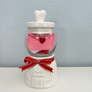 May include: White ceramic jar with a heart-shaped lid, filled with pink felt envelopes with red hearts. The jar has a red and white ribbon around the base and the text "5&cent;" on the front.
