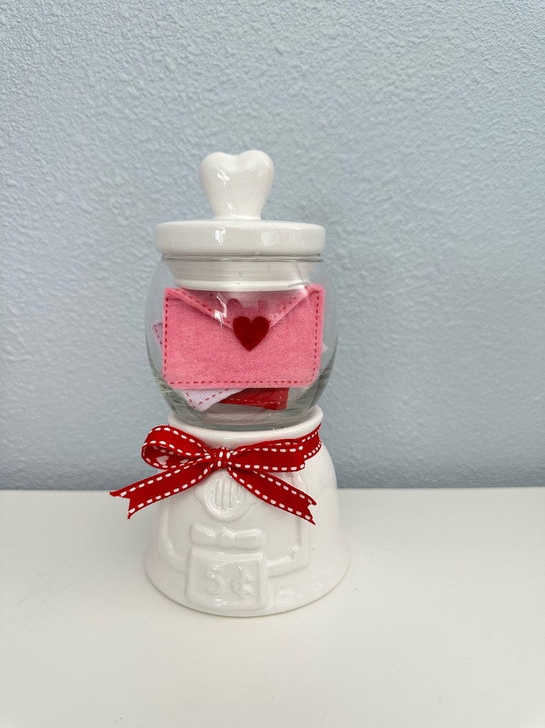 May include: White ceramic jar with a heart-shaped lid, filled with pink felt envelopes with red hearts. The jar has a red and white ribbon around the base and the text "5&cent;" on the front.