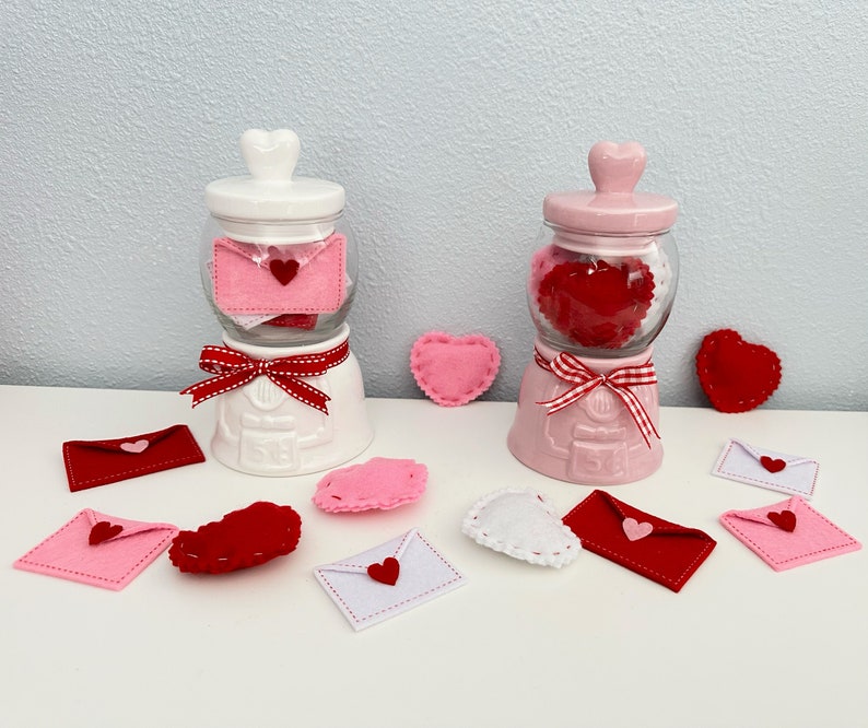 May include: Two white ceramic jars with lids shaped like hearts, filled with felt hearts. The jars are decorated with red and white ribbon. The jars are surrounded by felt hearts in various colors and shapes.