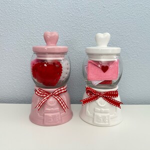 May include: Two ceramic Valentine's Day gumball machines. The pink one is filled with red and white felt hearts. The white one is filled with pink and white felt envelopes with a red felt heart on each envelope.