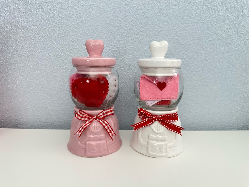 May include: Two ceramic Valentine's Day gumball machines. The pink one is filled with red and white felt hearts. The white one is filled with pink and white felt envelopes with a red felt heart on each envelope.