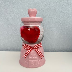 May include: Pink ceramic jar with a heart-shaped lid, filled with red and white felt hearts. The jar has a red and white gingham ribbon tied around the base and the number "54" printed on the front.