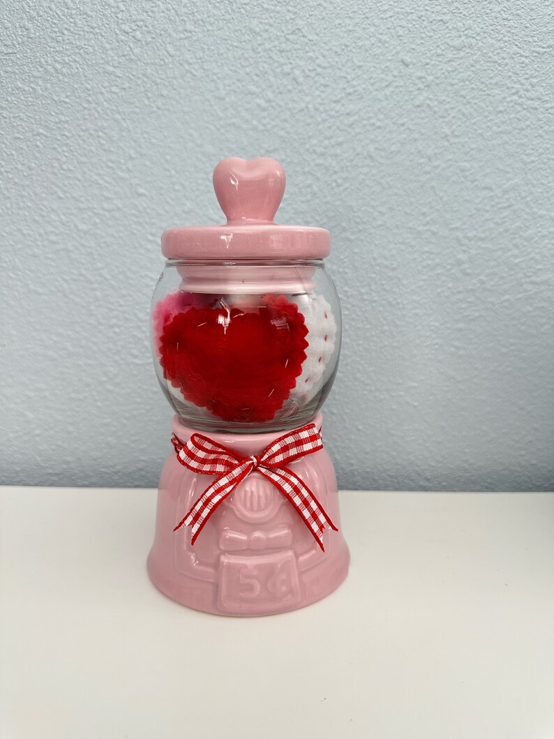 May include: Pink ceramic jar with a heart-shaped lid, filled with red and white felt hearts. The jar has a red and white gingham ribbon tied around the base and the number "54" printed on the front.
