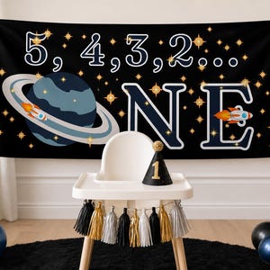 May include: A black banner with white numbers and the word "ONE" in large letters, decorated with a planet and a rocket ship. A high chair with a black and gold party hat and tassels is in front of the banner. Balloons in black, silver, and blue are on either side.