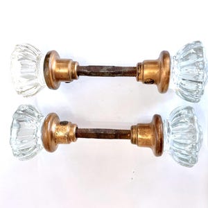 May include: Two vintage clear glass door knobs with a ribbed design. The knobs have brass-colored metal bases and are connected by a rusty metal rod. The knobs are likely from an antique door.