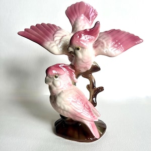 May include: A glossy ceramic figurine featuring two pink and white cockatoos perched on a brown branch. One bird is positioned lower with folded wings, while the other is perched above with its wings fully spread, showcasing pink-tipped feathers.