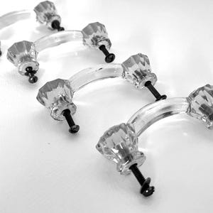 May include: A set of four clear, faceted glass drawer pulls arranged in a diagonal line. Each handle features a curved, transparent bar with a multi-faceted knob at each end, secured by a black metal screw extending from the base of each knob.