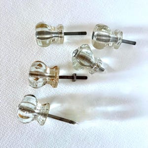 May include: Five clear glass cabinet knobs with a fluted, ribbed design and a rounded shape. Each knob features a central metal bolt extending from the back, designed for attachment to furniture drawers or doors, showcasing a transparent finish.