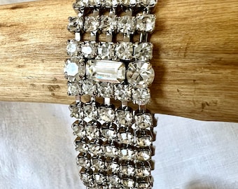 May include: A vintage rhinestone bracelet with multiple rows of clear, sparkling crystals. The bracelet is silver-toned and features a mix of round and rectangular stones, creating a glamorous accessory. The bracelet is displayed on a wooden surface.